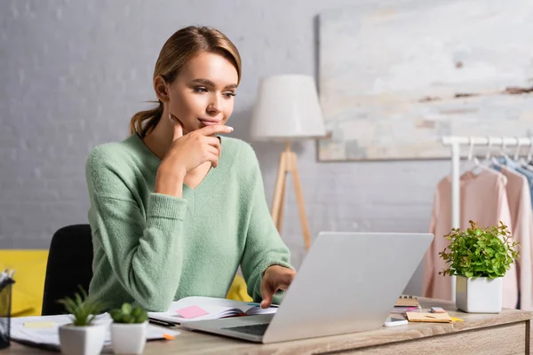 Freelance sorridente che usa il computer portatile mentre lavora vicino a notebook e piante in primo piano sfocato a casa — Foto stock