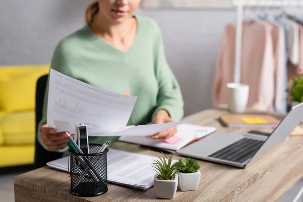 Plants and stationery on table near freelancer holding papers and laptop on blurred background — Stock Photo