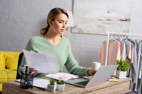 Rubia freelancer sosteniendo documentos mientras usa el ordenador portátil cerca de la taza y las plantas en primer plano borroso - foto de stock