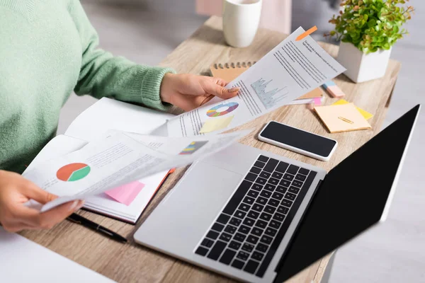 Vista recortada del teletrabajador sosteniendo documentos con gráficos cerca de dispositivos con pantalla en blanco en primer plano borroso - foto de stock