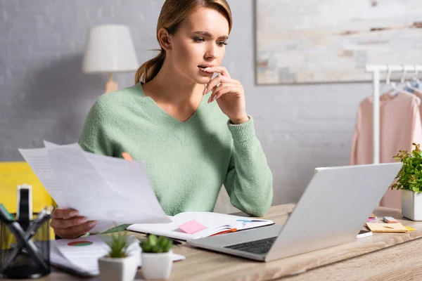 Nachdenklicher Freiberufler blickt auf Laptop neben Zeitungen und Notizbuch im verschwommenen Vordergrund — Stockfoto