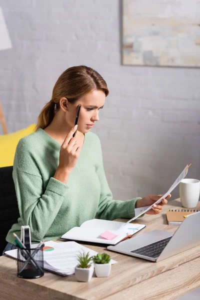 Pensativo freelancer celebración de la pluma mientras se trabaja con papeles cerca de la computadora portátil y plantas en primer plano borrosa - foto de stock