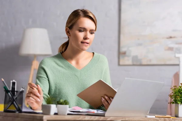 Freelancer pensativo segurando notebook perto de laptop e papelaria em primeiro plano desfocado — Fotografia de Stock