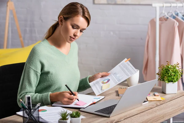 Freelancer escribiendo en un cuaderno mientras sostiene el documento con gráficos cerca de la computadora portátil en la mesa - foto de stock