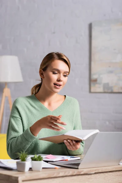 Smiling freelancer holding notebook during video call on laptop on blurred foreground — Stock Photo