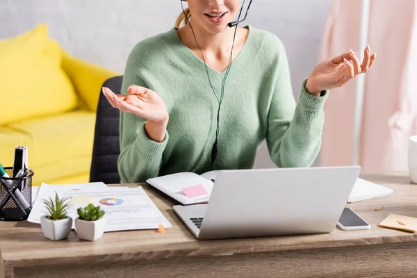 Cropped view of freelancer using headset during video call on laptop near papers on blurred foreground — Stock Photo