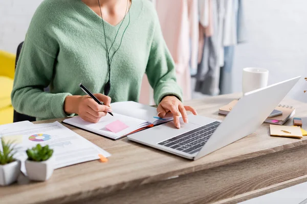 Vista recortada del freelancer usando el ordenador portátil y la escritura en el cuaderno cerca de papeles en primer plano borroso - foto de stock