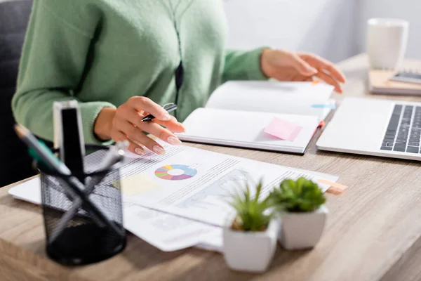 Vue recadrée du stylo freelance tenant près des papiers avec des graphiques, ordinateur portable et ordinateur portable sur fond flou — Photo de stock