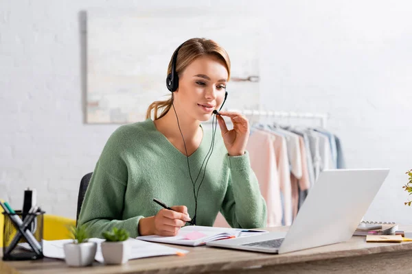 Freelance souriant à l'aide d'un casque et tenant un stylo près du portable pendant le chat vidéo sur ordinateur portable sur le premier plan flou — Photo de stock