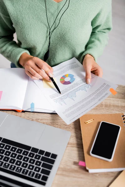 Vista recortada del freelancer sosteniendo pluma y documento con gráficos cerca de dispositivos y cuadernos en primer plano borroso - foto de stock