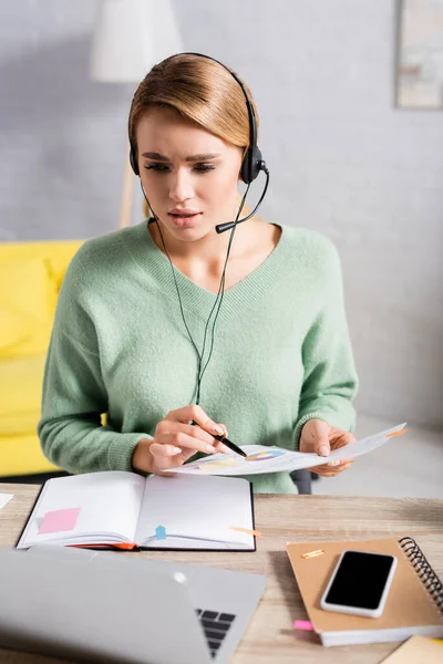 Freelancer in headset holding pen and document during video chat on laptop on blurred foreground — Stock Photo