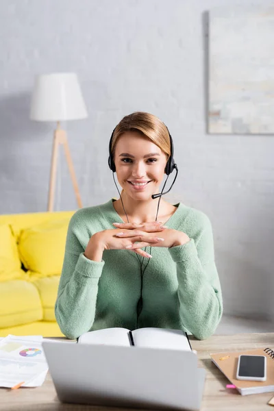 Smiling freelancer in headset looking at camera near notebooks, papers and devices on blurred foreground at home — Stock Photo