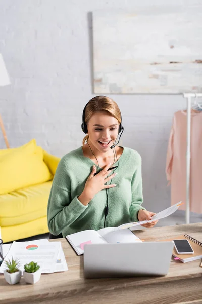 Freelancer sorridente em headset segurando caneta e papéis durante o bate-papo por vídeo no laptop em primeiro plano turvo — Fotografia de Stock