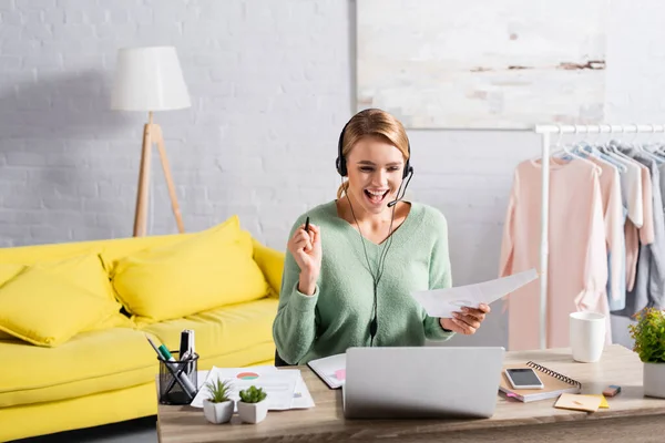 Freelancer alegre en la celebración de auriculares pluma y papel durante la videollamada en el ordenador portátil en casa - foto de stock