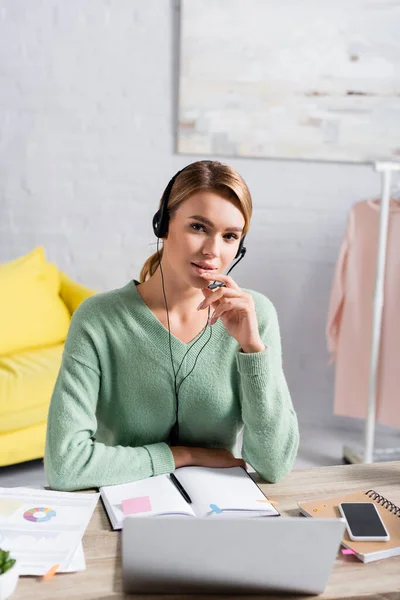 Blonde freelancer in headset looking at camera near notebooks, papers and devices on blurred foreground at home — Stock Photo