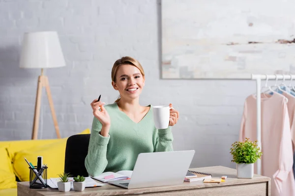 Cheerful freelancer holding cup and pen while working with papers and laptop on blurred foreground at home — Stock Photo