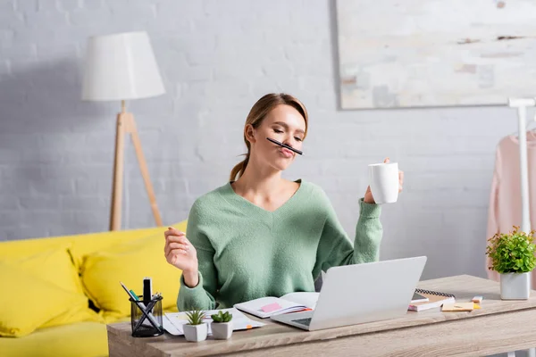 Freiberuflerin hält Tasse und Stift in Lippennähe, während sie zu Hause mit Papieren und Laptop arbeitet — Stockfoto