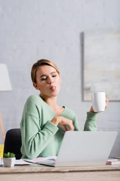 Young freelancer sticking out tongue and holding cup during video call on laptop on blurred foreground — Stock Photo