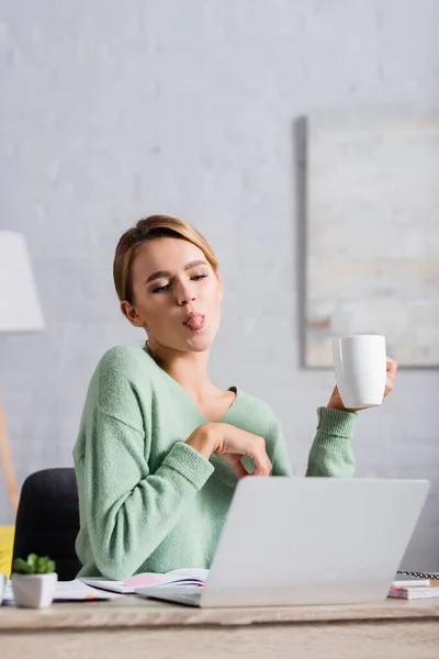 Junge Freiberuflerin streckt Zunge und Becher während Videoanruf am Laptop auf verschwommenem Vordergrund aus — Stockfoto