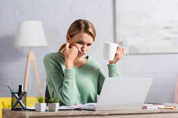 Skeptical freelancer holding cup while looking at laptop on blurred foreground on table — Stock Photo