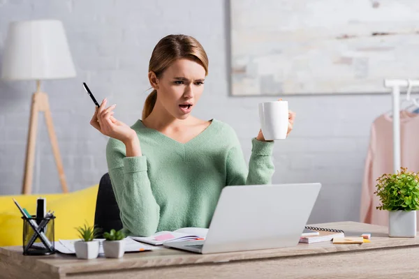 Confused freelancer holding pen and cup while looking at laptop near notebooks on blurred foreground — Stock Photo