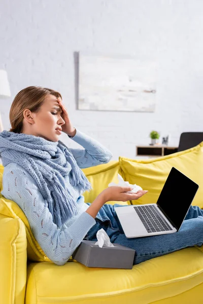 Sick woman with scarf holding napkin and looking at laptop with blank screen on couch — Stock Photo