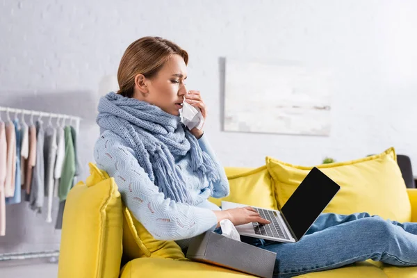 Ill freelancer in scarf holding napkin near nose while using laptop on sofa — Stock Photo