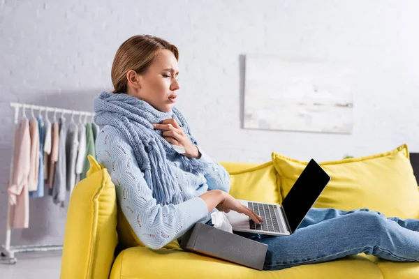 Mujer enferma sosteniendo la servilleta mientras usa el ordenador portátil en el sofá - foto de stock