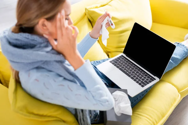 Laptop com tela em branco perto de mulher doente com termômetro e guardanapos em primeiro plano desfocado no sofá — Fotografia de Stock