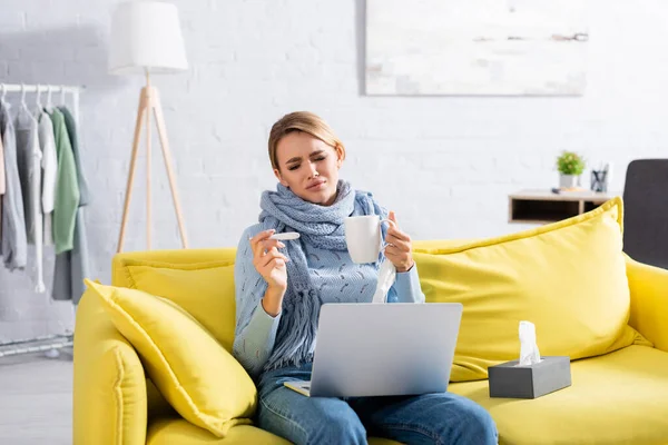 Femme malade tenant un thermomètre et une tasse près des serviettes et de l'ordinateur portable sur le canapé — Photo de stock