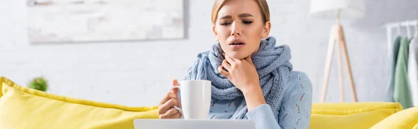 Donna malata in sciarpa che tiene la tazza vicino al computer portatile a casa, banner — Foto stock