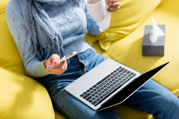 Cropped view of freelancer holding thermometer and cup near laptop and napkins on blurred background — Stock Photo