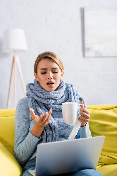 Enfermo freelancer celebración de la taza y tener videollamada en el ordenador portátil en primer plano borrosa — Stock Photo