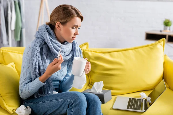 Enfermo freelancer sosteniendo taza y termómetro mientras mira el portátil cerca de servilletas en el sofá - foto de stock