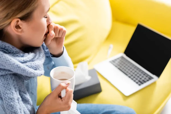 Femme malade toussant tout en tenant la tasse près de l'ordinateur portable et des serviettes sur fond flou — Photo de stock