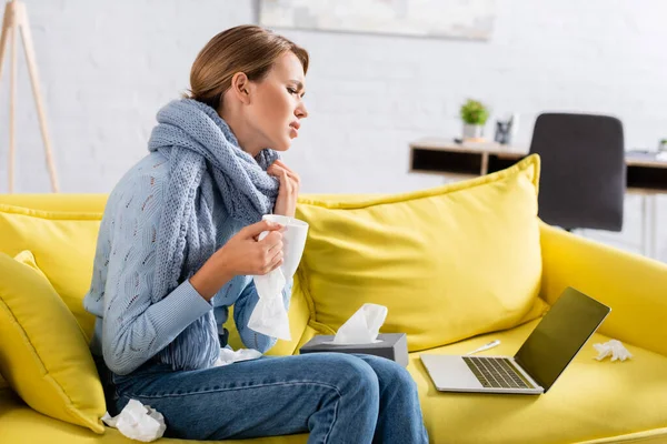 Kranker Freiberufler hält Tasse und Serviette in der Nähe von Laptop und Thermometer auf Couch — Stockfoto