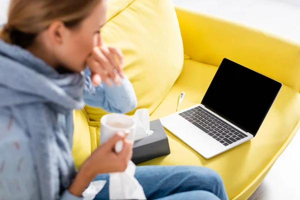 Ноутбук з порожнім екраном на дивані біля термометра і хвора жінка з чашкою на розмитому передньому плані — стокове фото
