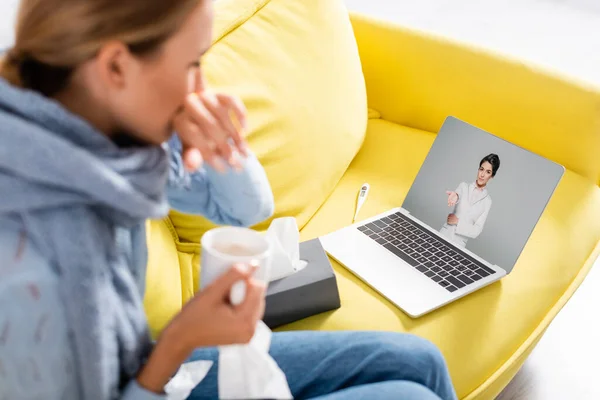 Doctor señalando con la mano en la pantalla de la computadora portátil cerca de la mujer enferma sosteniendo la taza durante la videollamada en primer plano borroso - foto de stock