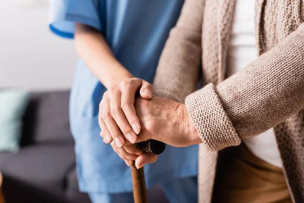 Vista parziale di infermiera toccare le mani della donna anziana in piedi con bastone da passeggio, sfondo sfocato — Foto stock