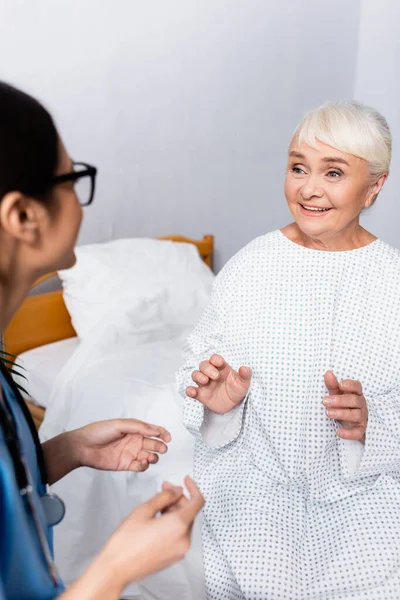 Счастливая пожилая женщина жестом во время разговора с медсестрой на размытом переднем плане — стоковое фото