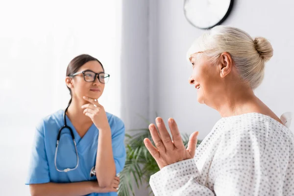 Jovem asiático enfermeira no óculos ouvindo idosos mulher falando e gestos no hospital, desfocado fundo — Fotografia de Stock
