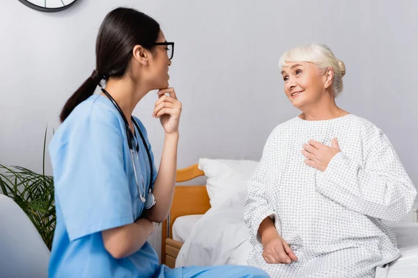 Веселая пожилая женщина держит руку на груди, разговаривая в больнице с азиатской медсестрой — стоковое фото