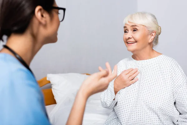 Positive elderly woman touching chest while talking to nurse gesturing on blurred foreground — Stock Photo