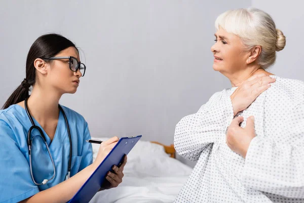 Young asian nurse writing on clipboard near senior woman suffering from pain in shoulder — Stock Photo