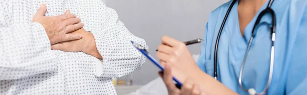 Vista recortada de la mujer mayor tocando el pecho mientras sufría de un ataque de jadeo cerca de la enfermera escribiendo en el portapapeles, borrosa primer plano, pancarta - foto de stock