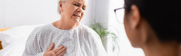 Mujer mayor tocando el pecho mientras sufre de dolor de corazón cerca de la enfermera en primer plano borrosa, bandera — Stock Photo