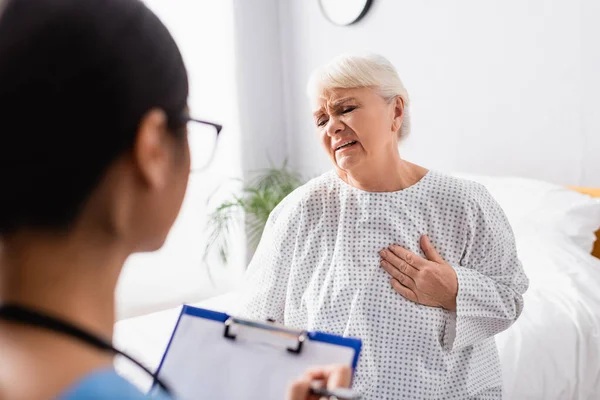 Seniorin berührt Brust, während sie unter Herzschmerzen in der Nähe von Krankenschwester leidet, die auf Klemmbrett schreibt, verschwommener Vordergrund — Stockfoto