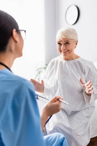 Happy elderly woman smiling and gesturing while talking to nurse on blurred foreground — Stock Photo