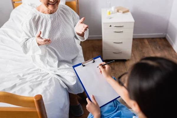 Overhead-Ansicht einer Krankenschwester, die auf Klemmbrett neben lächelnder und gestikulierender Seniorin schreibt, verschwommener Vordergrund — Stockfoto