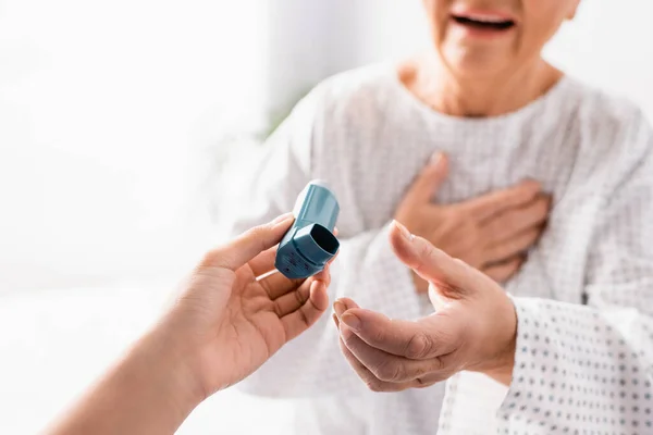 Vista recortada de la enfermera que da inhalador a una mujer anciana que sufre de un ataque de asma en un fondo borroso - foto de stock